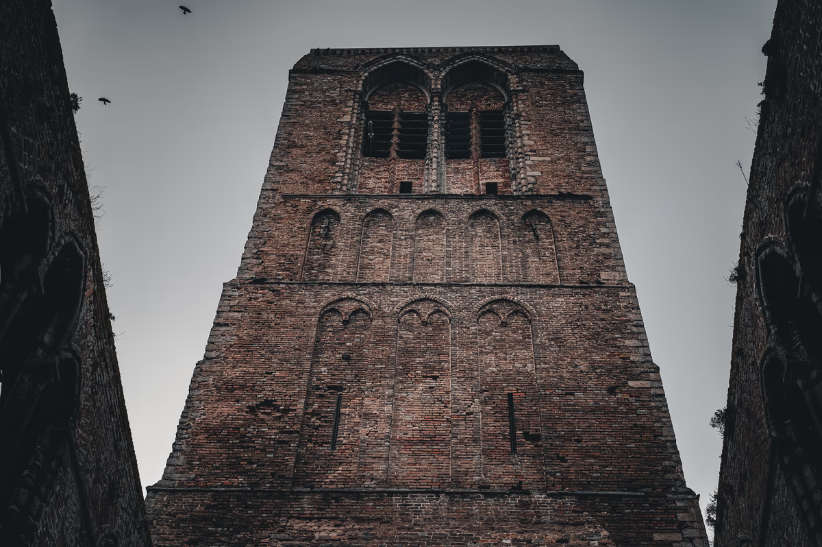 Low-angle shot of a weathered, medieval brick tower with arched windows and bird silhouettes against a gray sky.