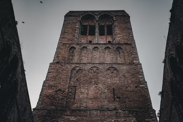 Low-angle shot of a weathered, medieval brick tower with arched windows and bird silhouettes against a gray sky.