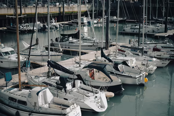 High-angle view of several white sailboats and motorboats docked side-by-side at a harbor pier on calm, teal-toned water.