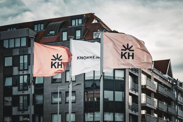 Three flags with the 'KH' logo and 'Knokke' text flutter in the wind against the backdrop of a modern apartment building under a cloudy sky.