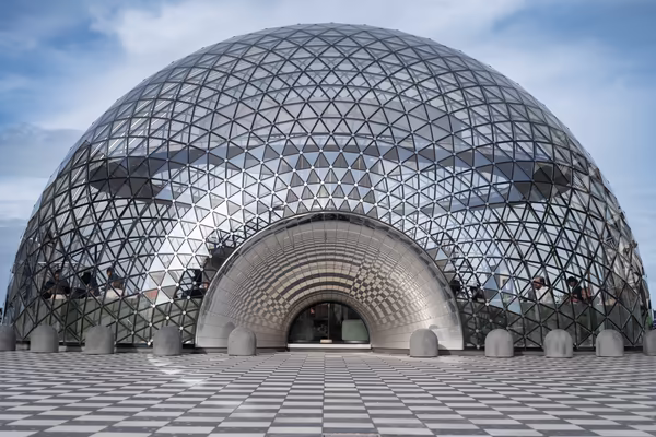 A symmetrical, low-angle shot of a large geodesic glass dome structure with a metallic arched entrance, situated on a black and white checkered pavement under a clear sky.