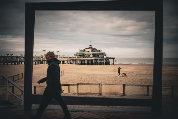 A wide-angle shot through a large rectangular wooden frame looking onto a beach. In the background, a grand pier building stands over the sand, while a person walks a dog. In the foreground, a blurred silhouette of a person passes by.