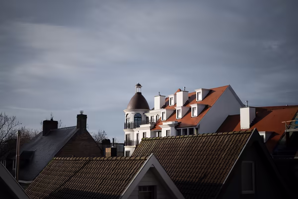 A low-angle shot of European-style rooftops under a cloudy sky, featuring a prominent white building with a dark dome and bright orange-tiled roofs catching the sunlight.