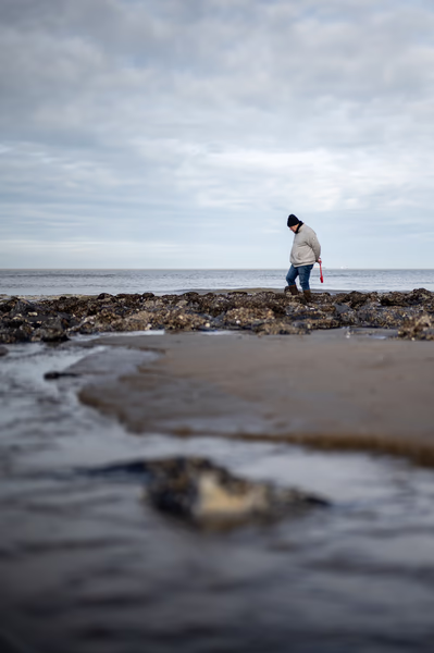 A person in a winter hat and jacket walks along a rocky, barnacle-covered shoreline at low tide under a cloudy sky.