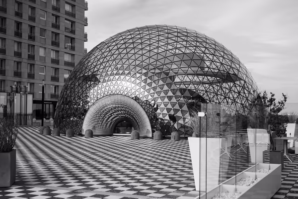 A high-contrast black and white photograph of a modern geodesic glass dome structure situated on a checkered plaza, featuring intricate patterns of light and shadow.